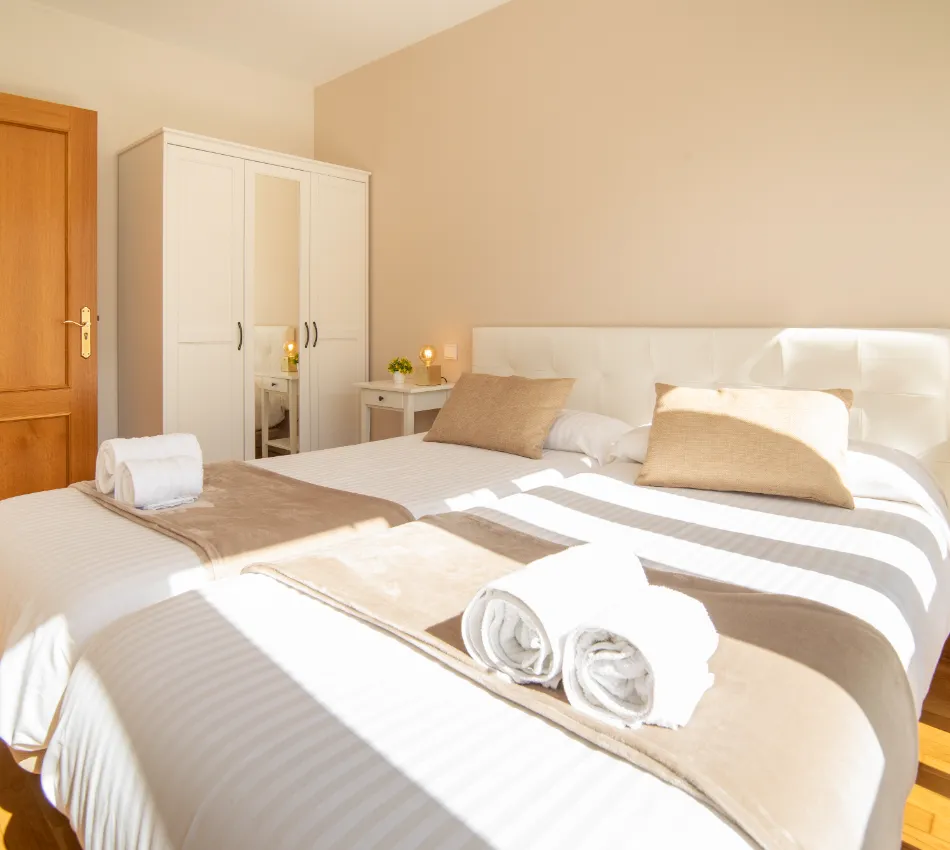 Habitaci&oacute;n luminosa con cama doble, dos almohadas, toallas enrolladas, armario y mesita de noche.