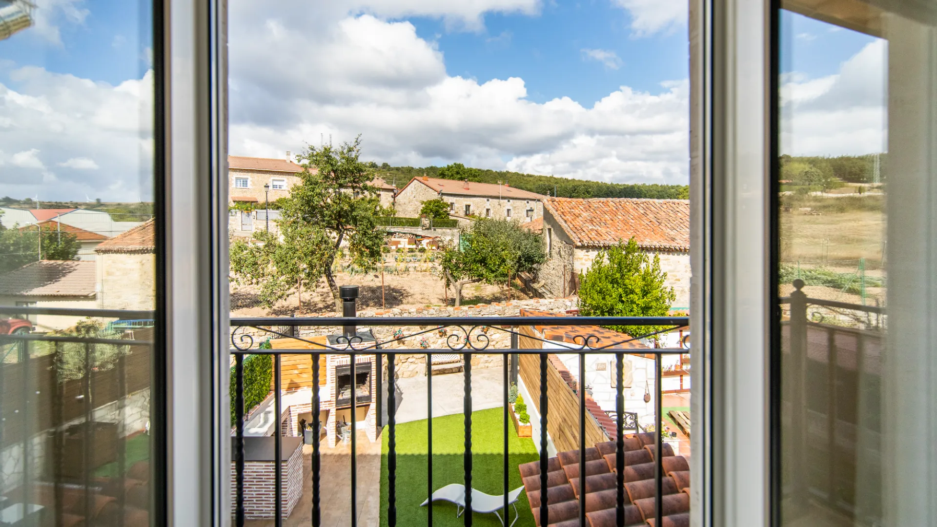 Vista desde una ventana a un jard&iacute;n con muebles y casas r&uacute;sticas al fondo bajo un cielo nublado.