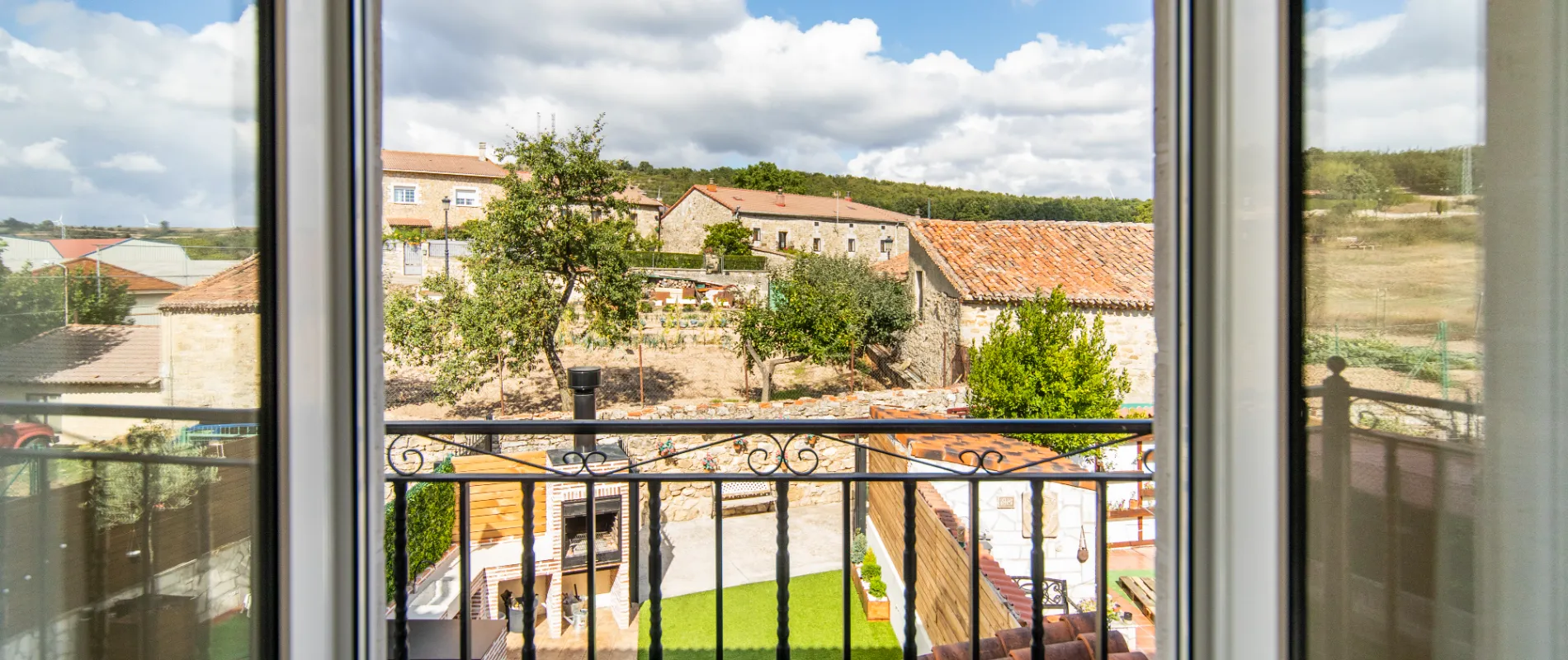 Vista desde una ventana a un jard&iacute;n con barbacoa y casas de campo al fondo, bajo un cielo azul.