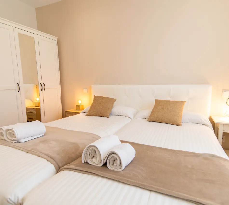 Habitaci&oacute;n con dos camas individuales, cojines marrones y toallas enrolladas sobre cobijas beige.