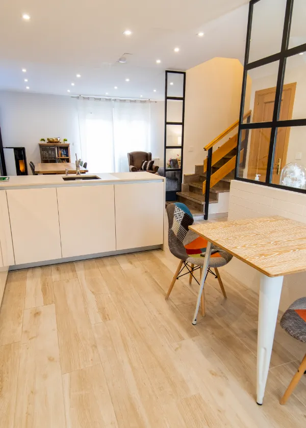 Sala moderna con escalera, mesa y sillas de dise&ntilde;o. Cocina abierta y librero al fondo.