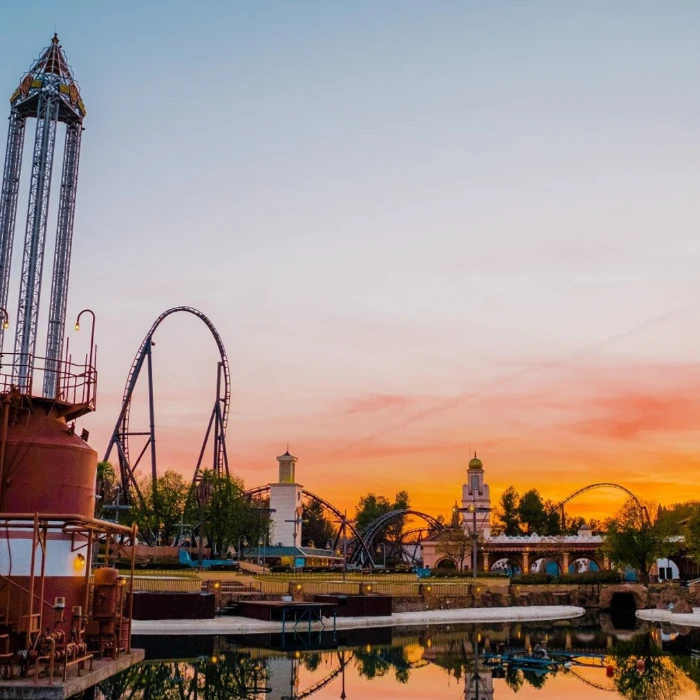 Parque de diversiones al atardecer con monta&ntilde;as rusas y un lago en primer plano.