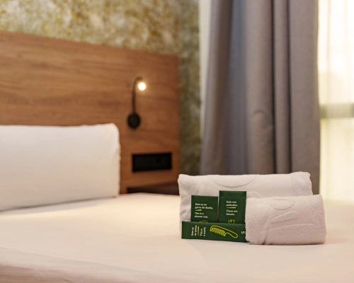 Habitaci&oacute;n de hotel con toallas dobladas sobre la cama y luz de lectura.