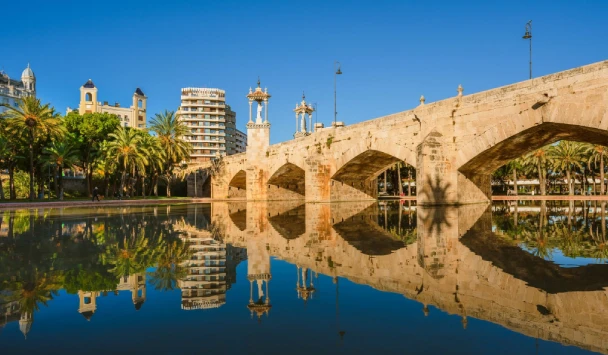 Puente de piedra reflejado en el agua, con edificios y palmeras al fondo.