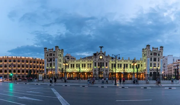 Estaci&oacute;n de tren iluminada al anochecer, con cielo nublado y gente alrededor.