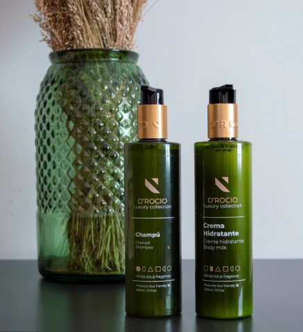 Botellas de champ&uacute; y crema hidratante D'ROCIO frente a un jarr&oacute;n verde.