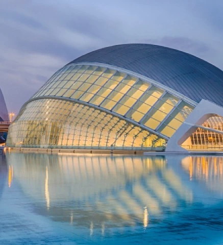 Edificio moderno con c&uacute;pula y reflejo en el agua al atardecer.