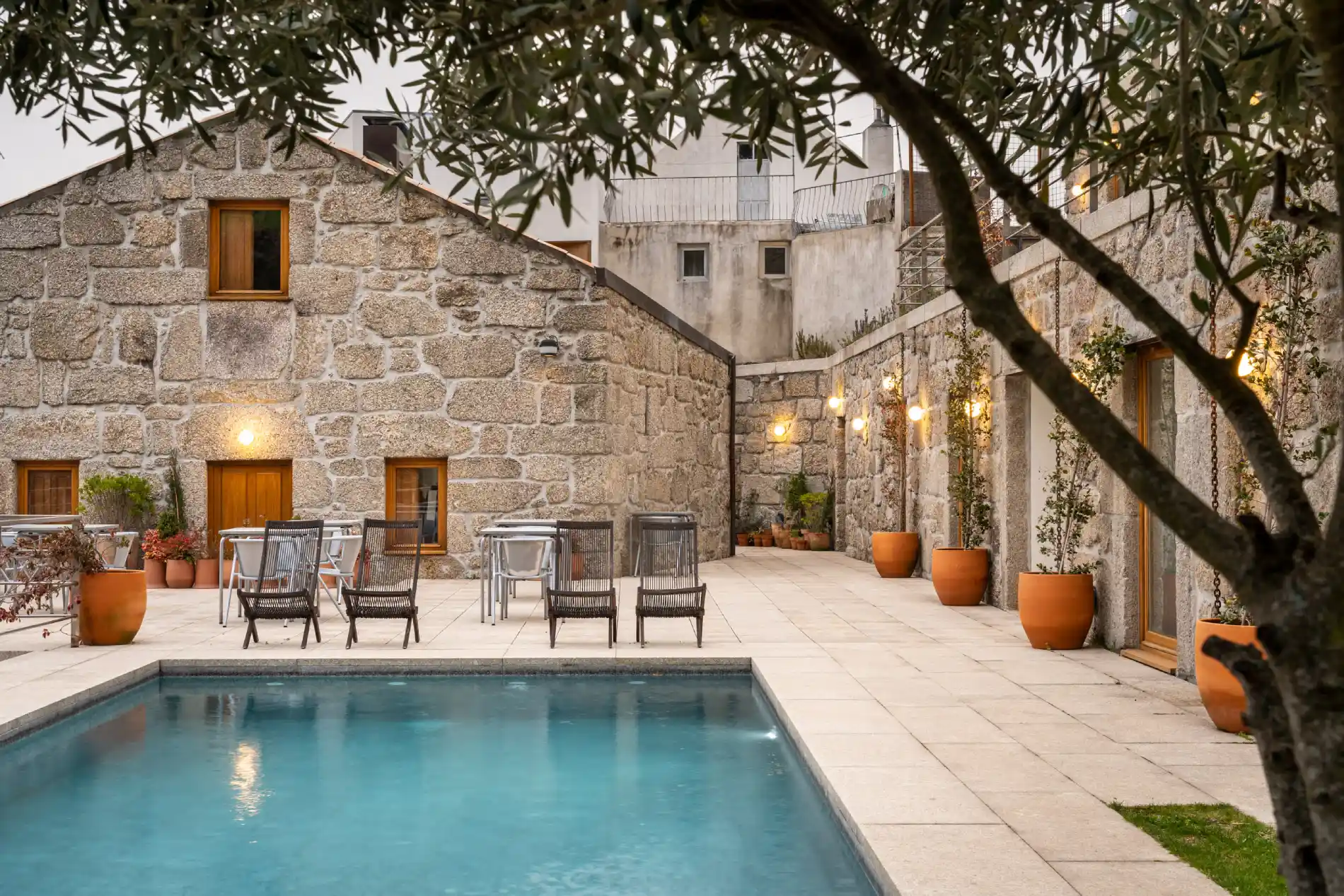 P&aacute;tio com piscina, cadeiras, mesas, e paredes de pedra sob luzes acesas e &aacute;rvore &agrave; frente.