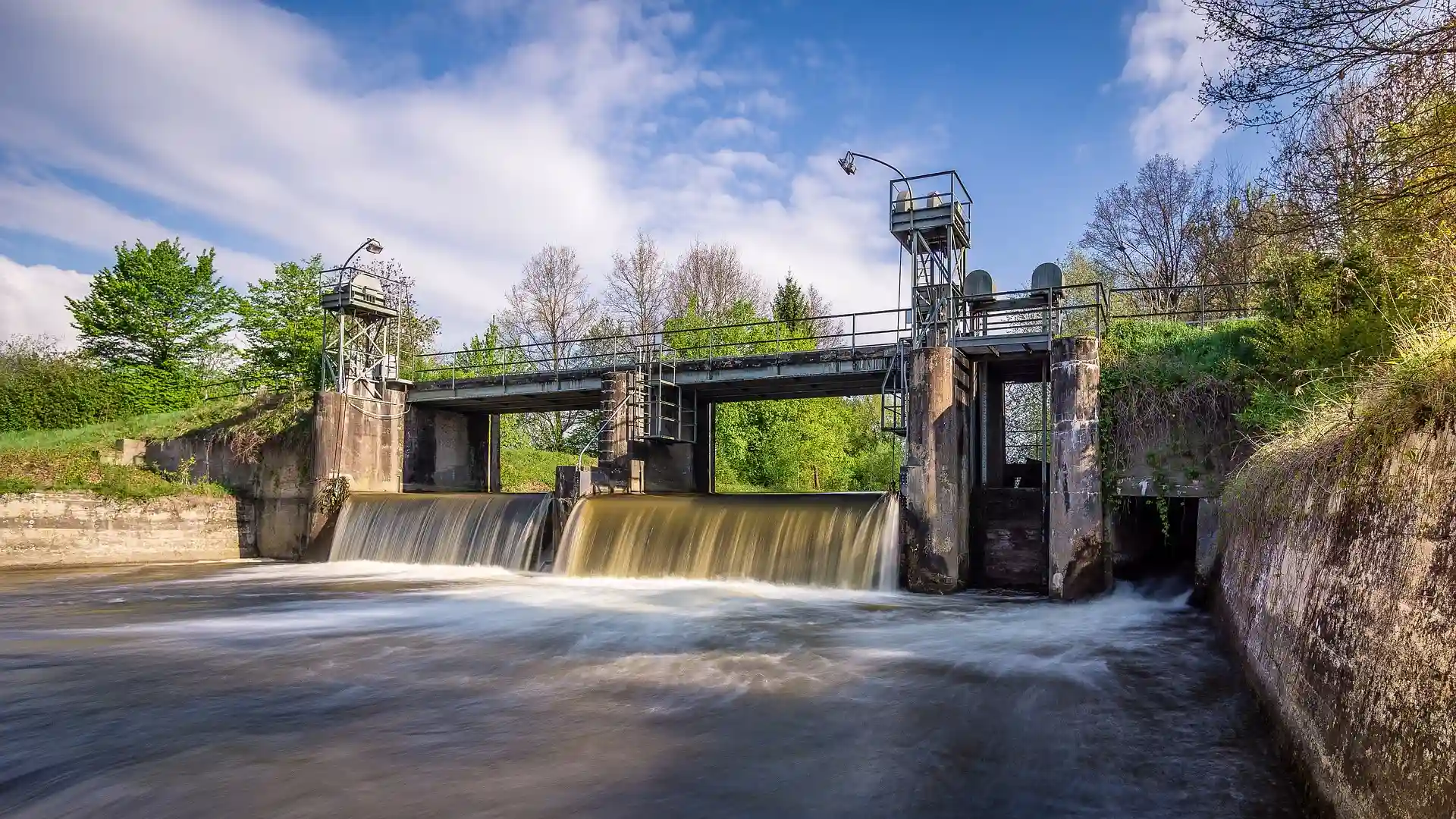Barragem de concreto com &aacute;gua fluindo, cercada por vegeta&ccedil;&atilde;o e c&eacute;u azul.