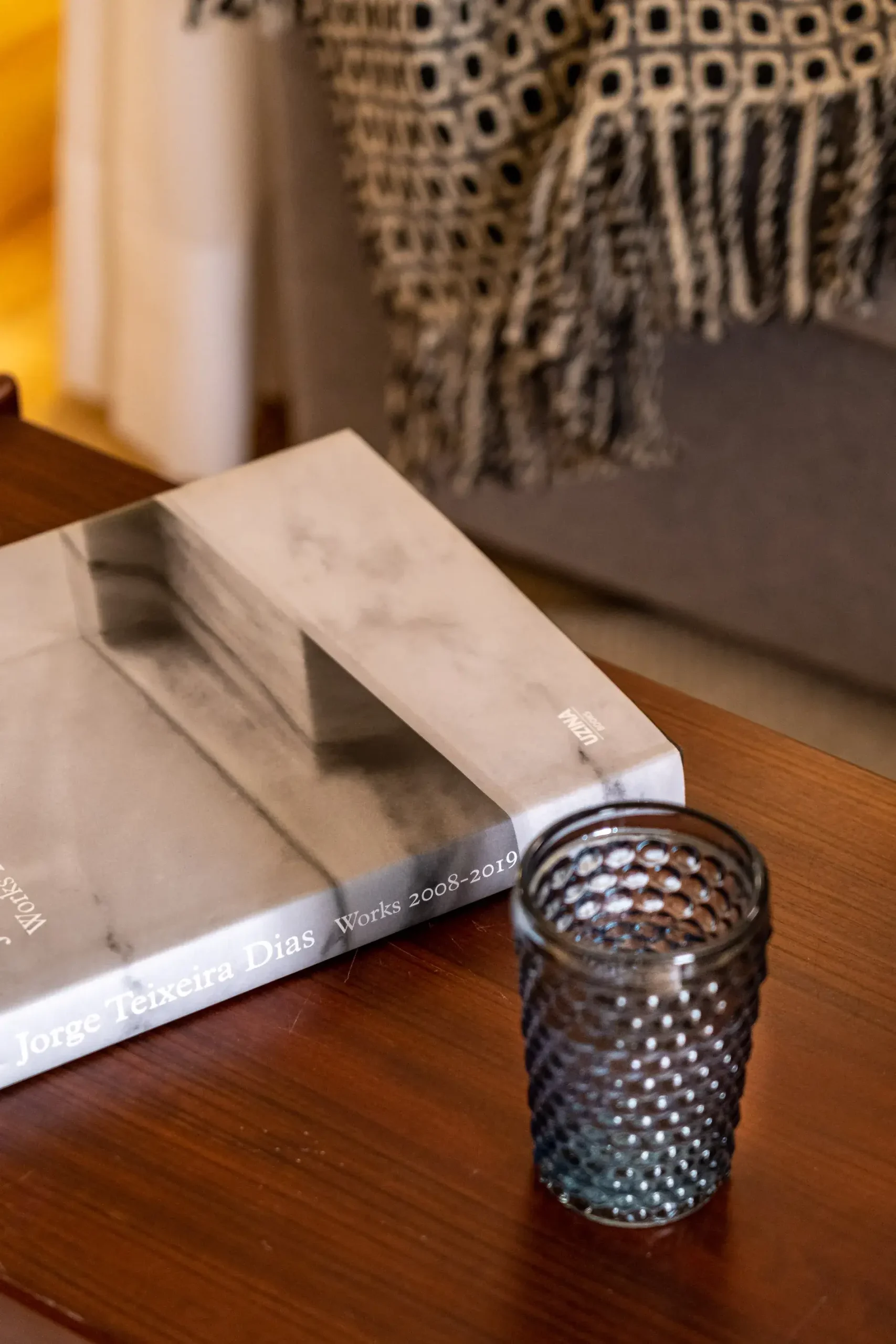 Livro sobre mesa de madeira ao lado de copo texturizado; manta com franjas ao fundo.