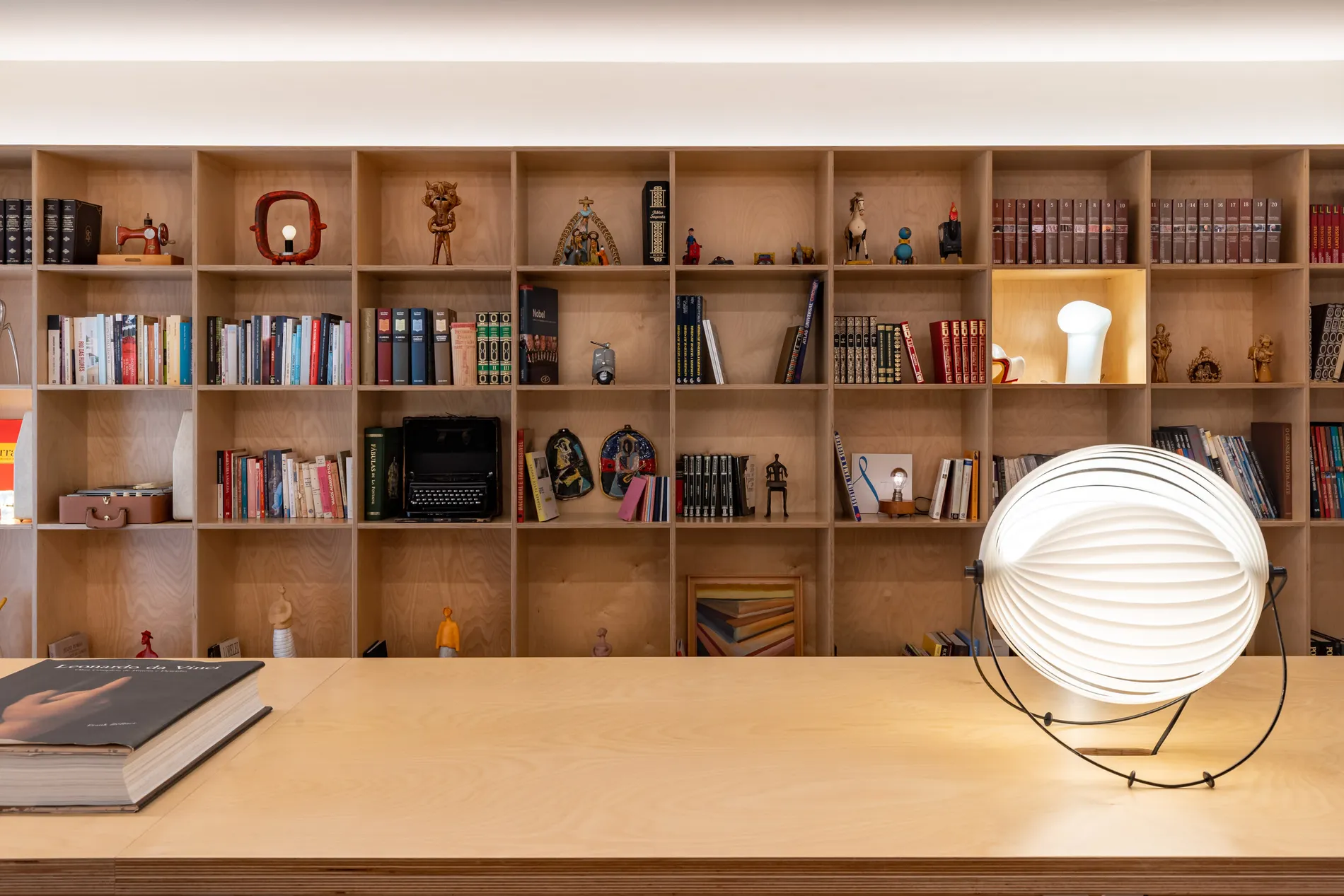 Estante de madeira com livros, objetos decorativos e uma lumin&aacute;ria moderna sobre a mesa.