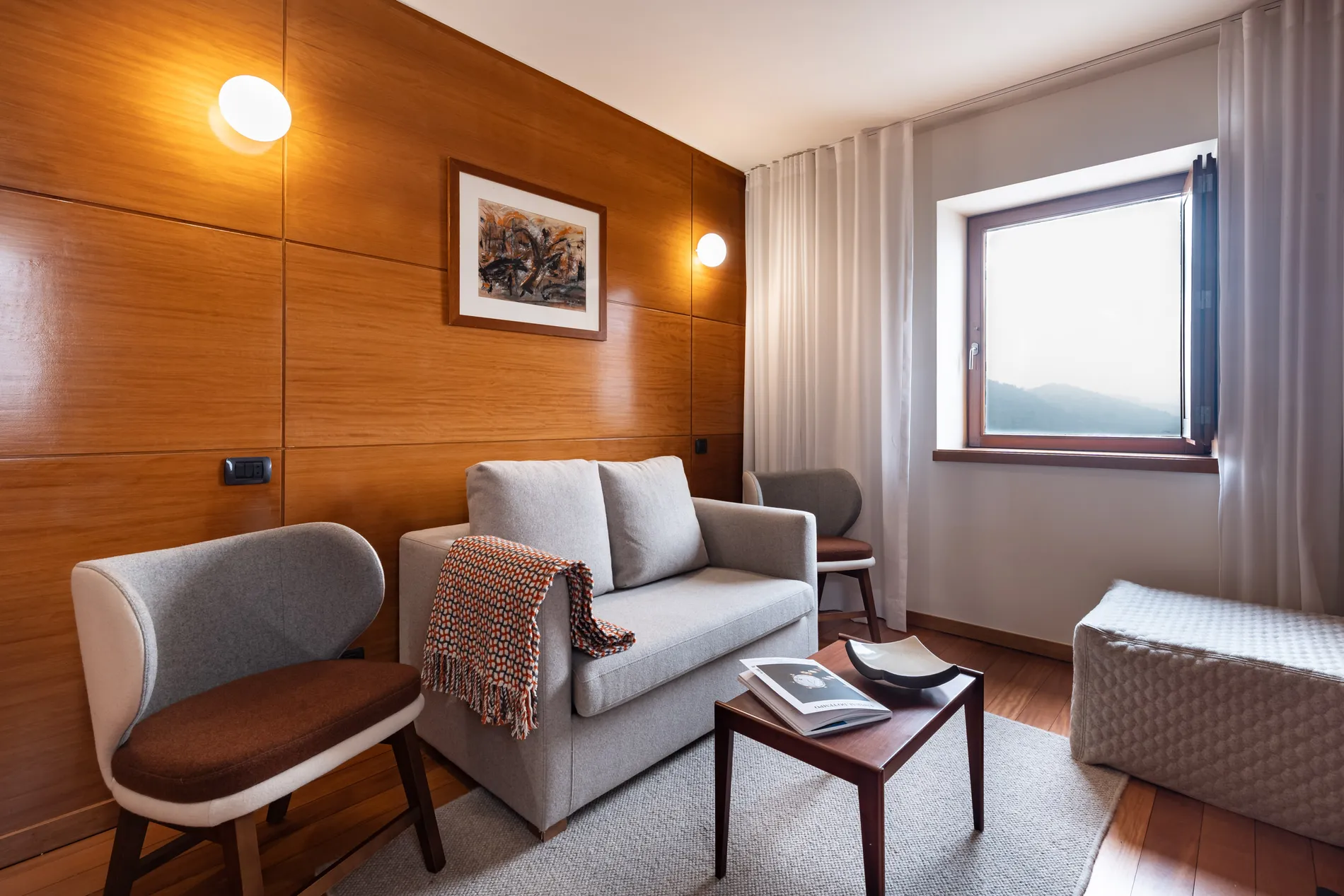 Sala com sof&aacute;, cadeiras, mesa de centro, luz suave e janela aberta com vista para montanhas.