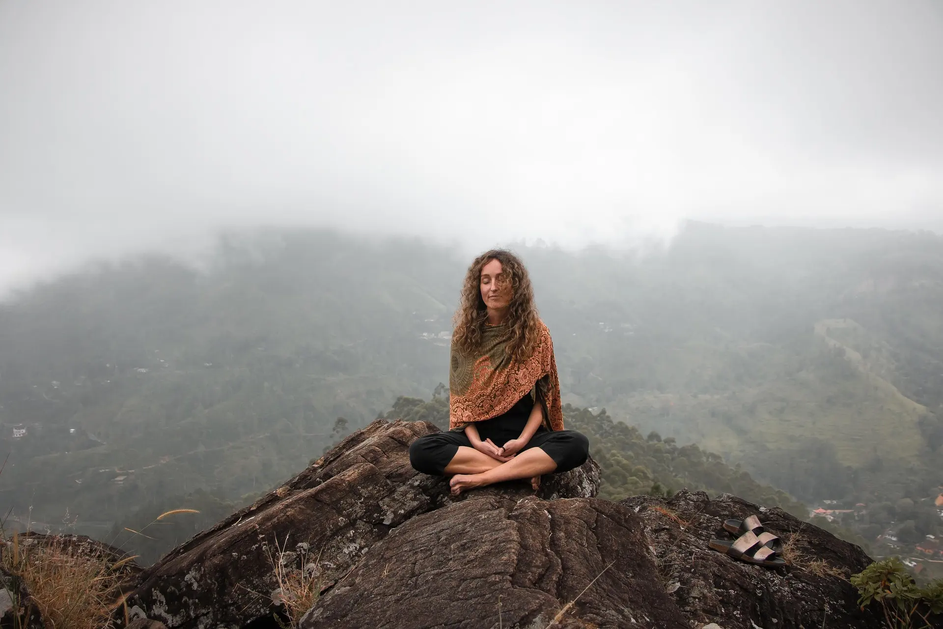 Mulher meditando em posi&ccedil;&atilde;o de l&oacute;tus sobre uma rocha em um local montanhoso e nublado.