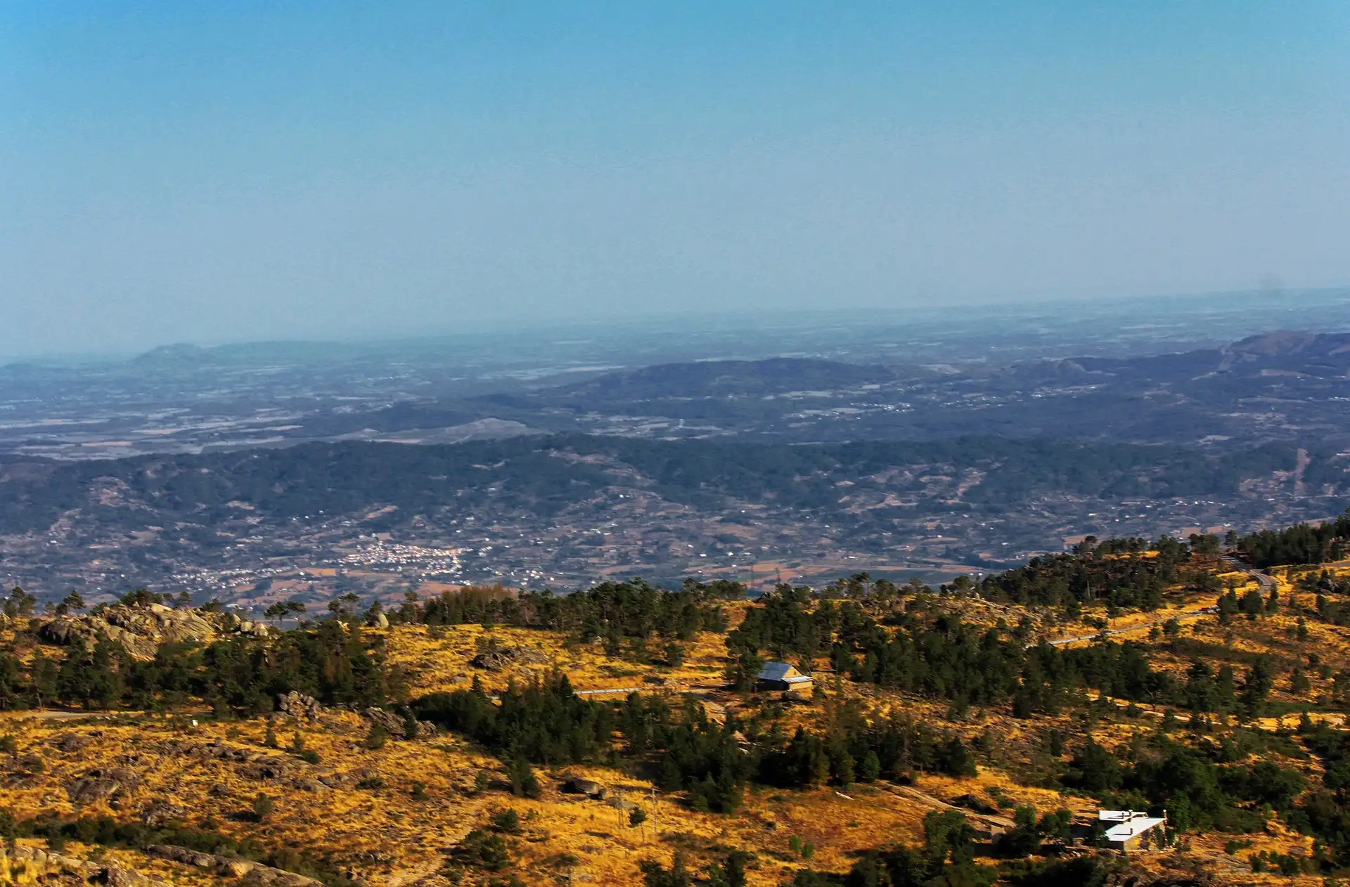 Vista panor&acirc;mica de montanhas e florestas sob um c&eacute;u claro, com casas ao longe.