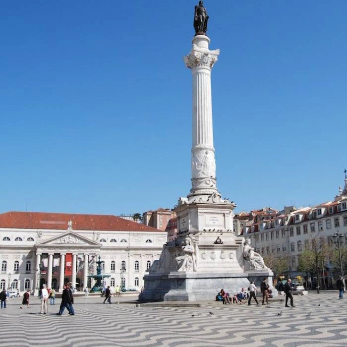 Pra&ccedil;a com coluna e est&aacute;tua central, pr&eacute;dios ao fundo, c&eacute;u azul.