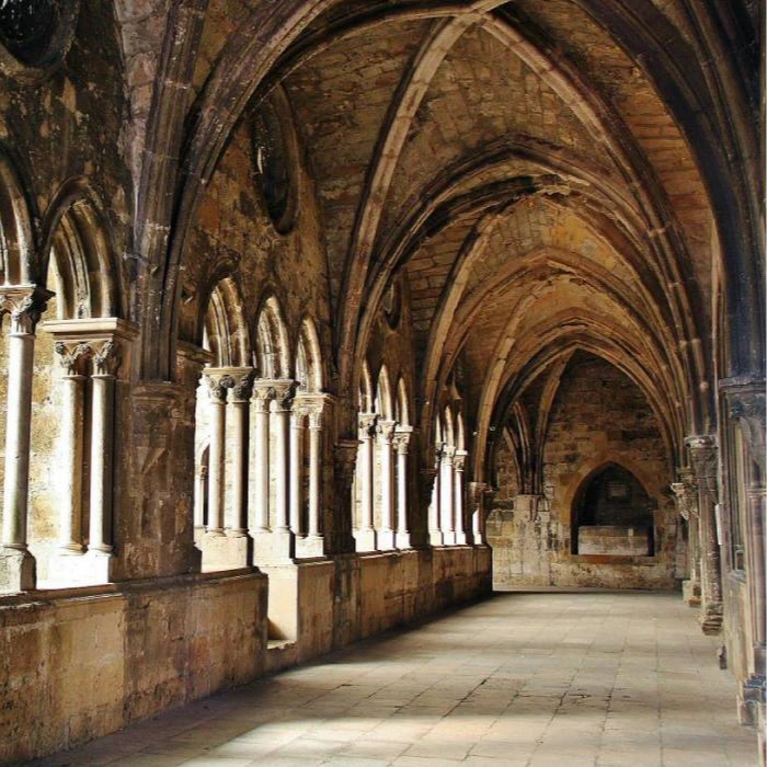 Corredor de pedra com colunas e arcos g&oacute;ticos em luz natural.