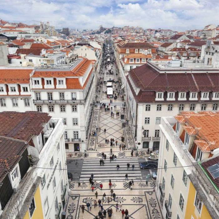 Rua movimentada com cal&ccedil;ada portuguesa entre pr&eacute;dios hist&oacute;ricos.