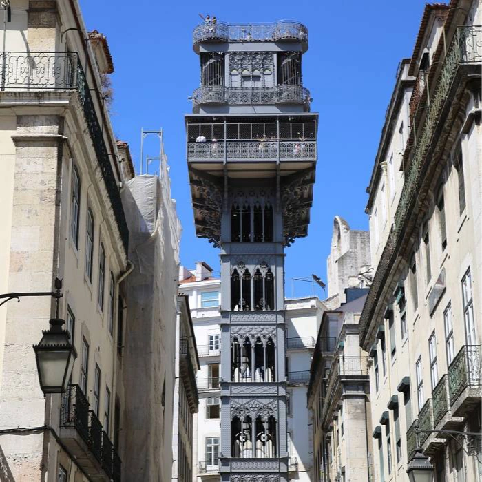 Elevador em estilo neog&oacute;tico entre pr&eacute;dios sob c&eacute;u azul.