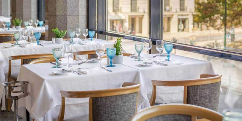 Restaurante elegante com mesas postas, janelas amplas e vista para a rua.