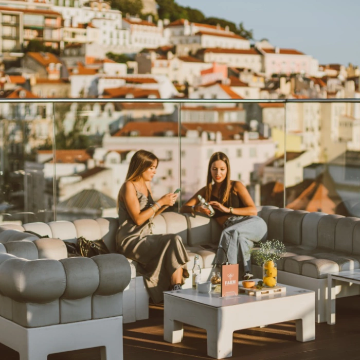 Duas mulheres conversam em um terra&ccedil;o com vista para a cidade ao fundo.