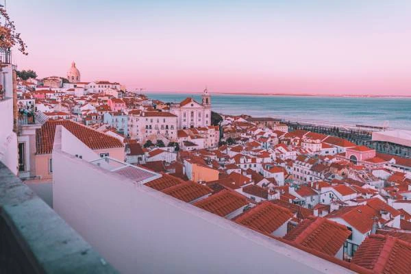 Casario de Lisboa sob c&eacute;u rosado ao amanhecer, com rio ao fundo.