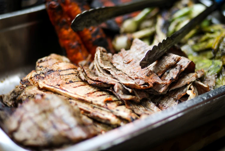 Carne asada al grill con pinzas en una bandeja, lista para servir.