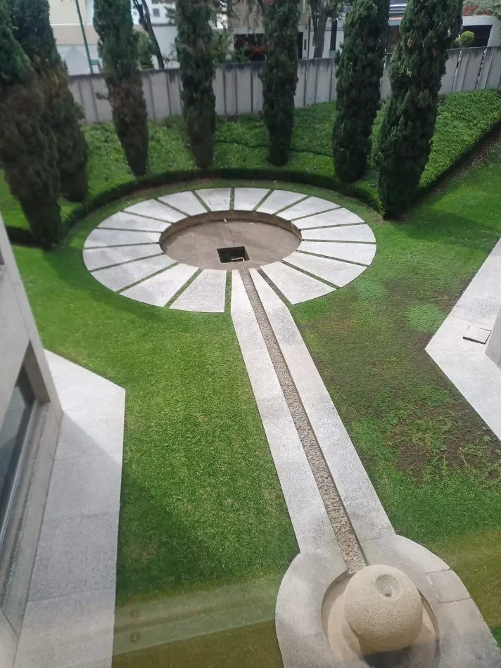 Jard&iacute;n circular con &aacute;rbolillos altos y pasillo central de piedra.