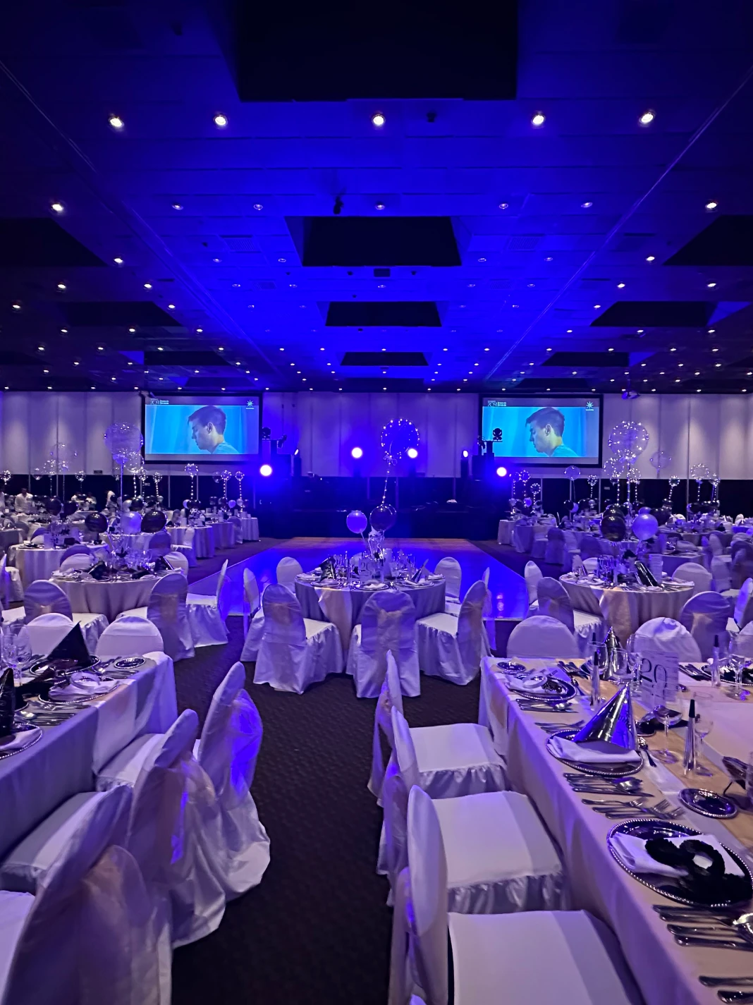 Sal&oacute;n de eventos decorado en tonos azules con mesas elegantes y pantallas grandes.