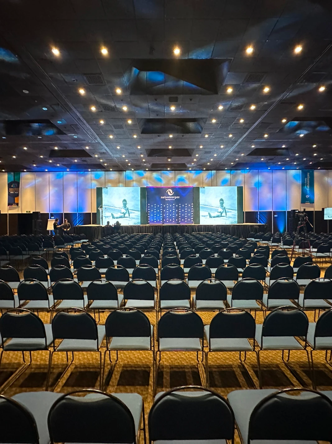 Auditorio vac&iacute;o con sillas y pantallas en el escenario iluminado.