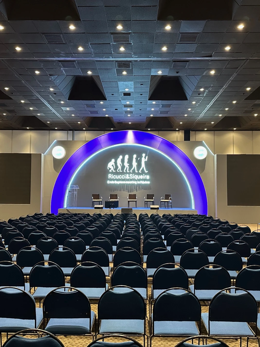 Auditorio con sillas azules y escenario iluminado con logotipo de evoluci&oacute;n humana.