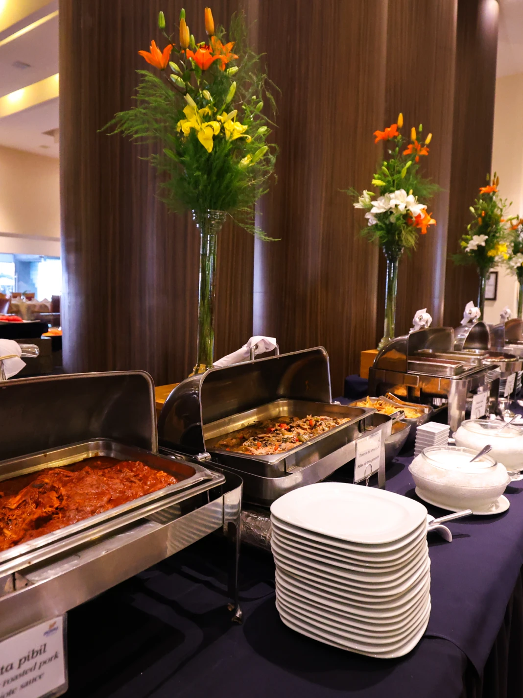 Bufet con platos listos para servir, rodeado de arreglos florales altos.