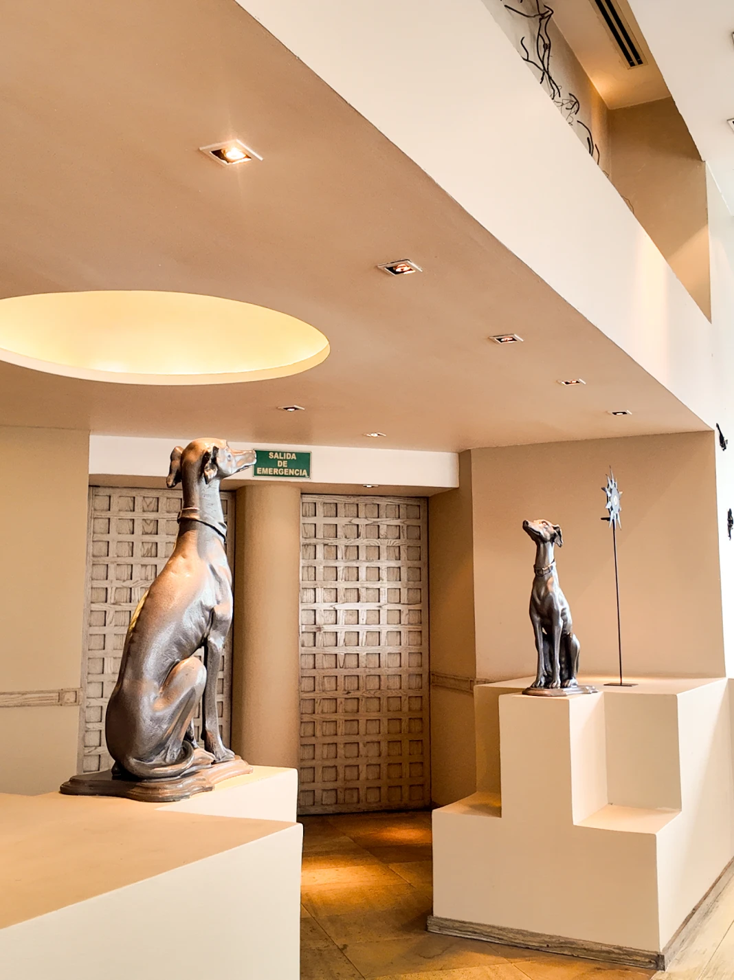 Esculturas de perros en un vest&iacute;bulo moderno con luz c&aacute;lida y se&ntilde;al de emergencia.