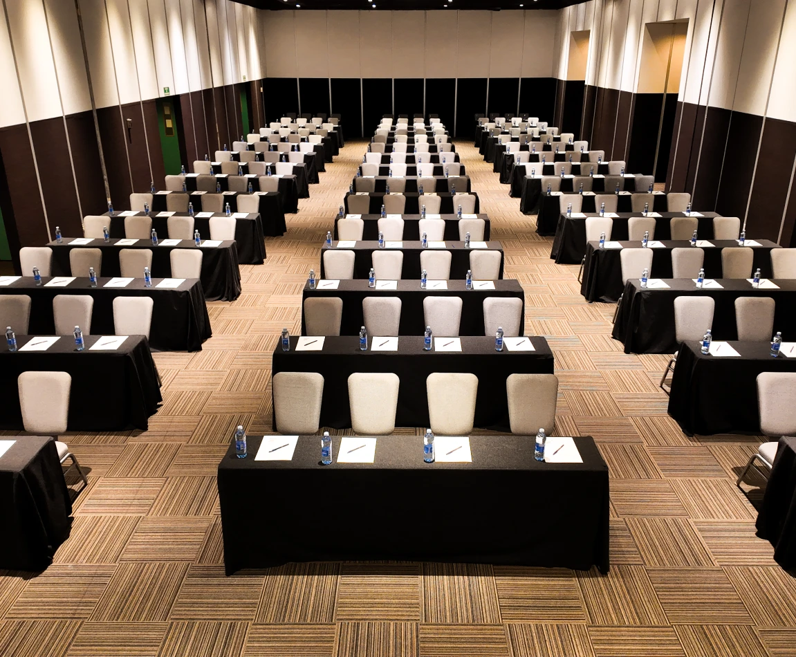 Sala de conferencias vac&iacute;a con mesas, sillas, botellas de agua y cuadernos.