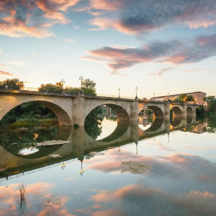 Puente de piedra sobre agua tranquila reflejando el cielo al atardecer.