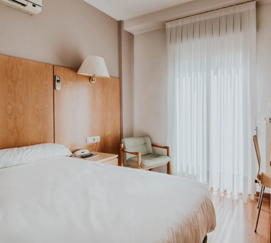 Habitaci&oacute;n de hotel con cama, silla y ventana con cortinas blancas.