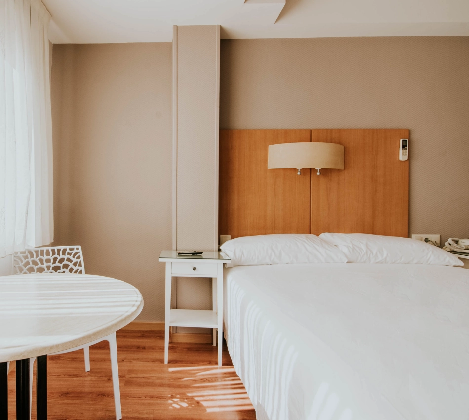 Habitaci&oacute;n de hotel moderna y luminosa con cama, mesa y silla junto a una ventana.