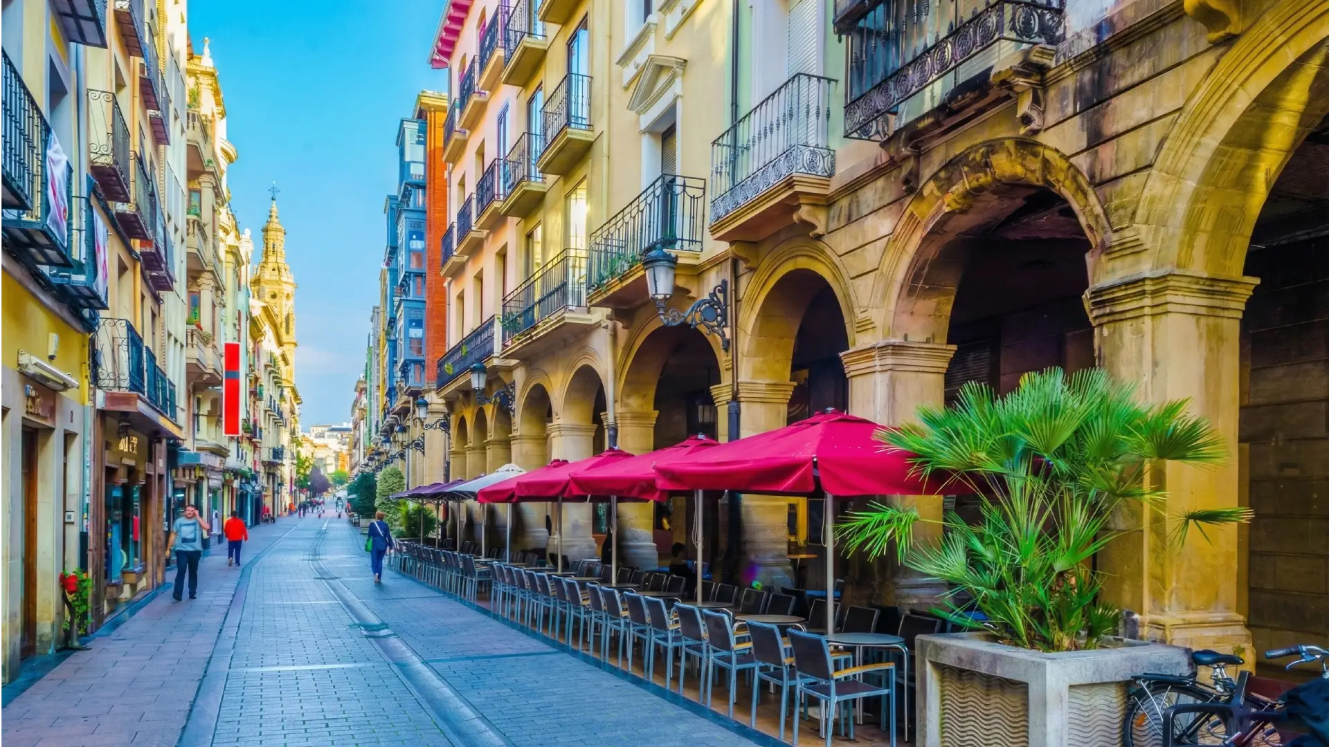 Calle con edificios, arcadas y sombrillas rojas junto a una torre al fondo.