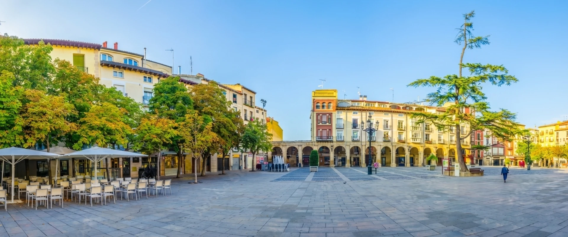 Plaza amplia con &aacute;rboles y edificios antiguos, cielo despejado.