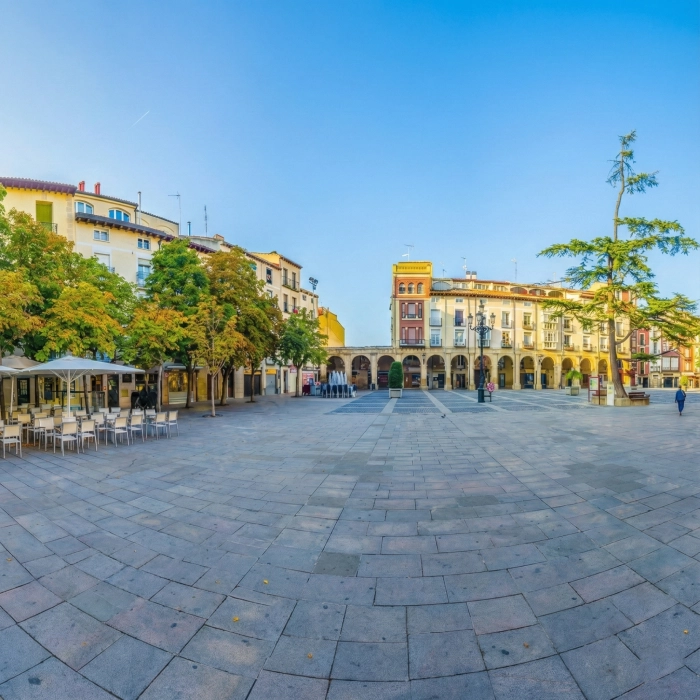 Plaza amplia con edificios hist&oacute;ricos y &aacute;rboles bajo un cielo azul despejado.