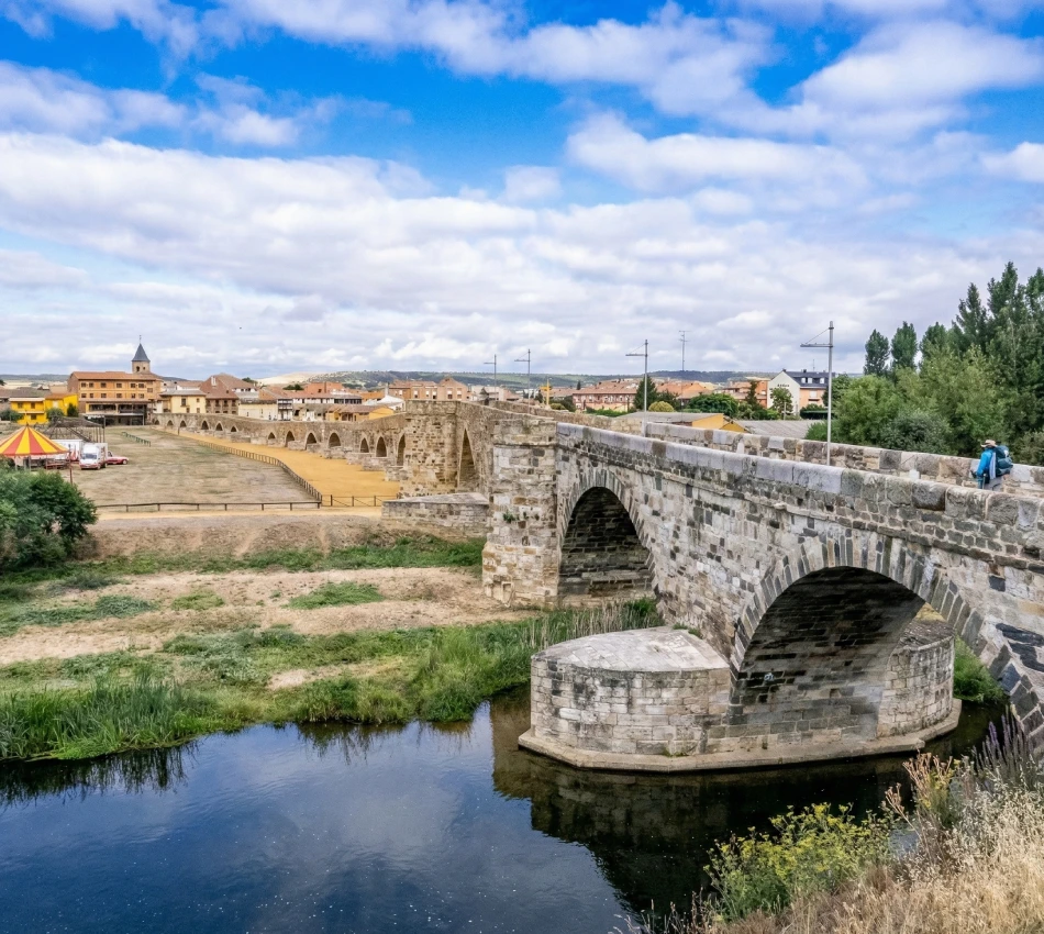 Puente de piedra junto a un r&iacute;o con un pueblo al fondo bajo un cielo azul.