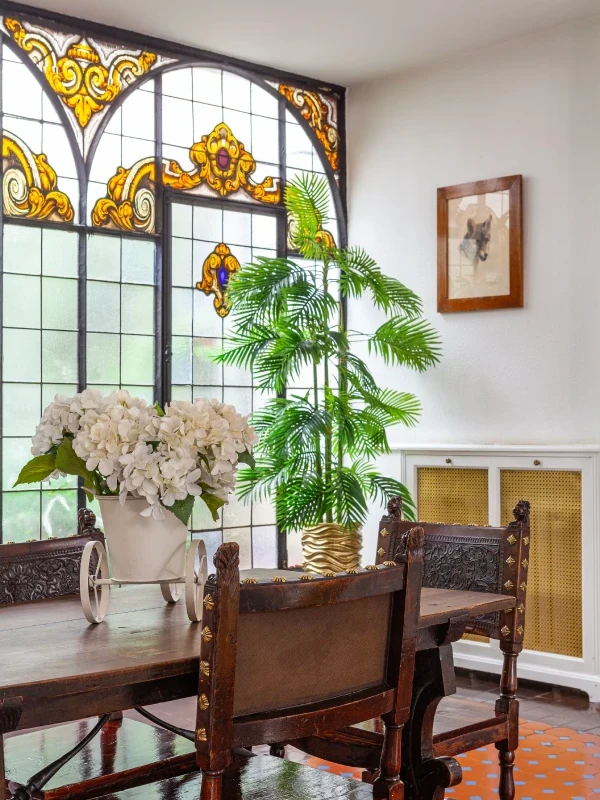 Comedor con vitrales coloridos, planta y flores blancas sobre la mesa.