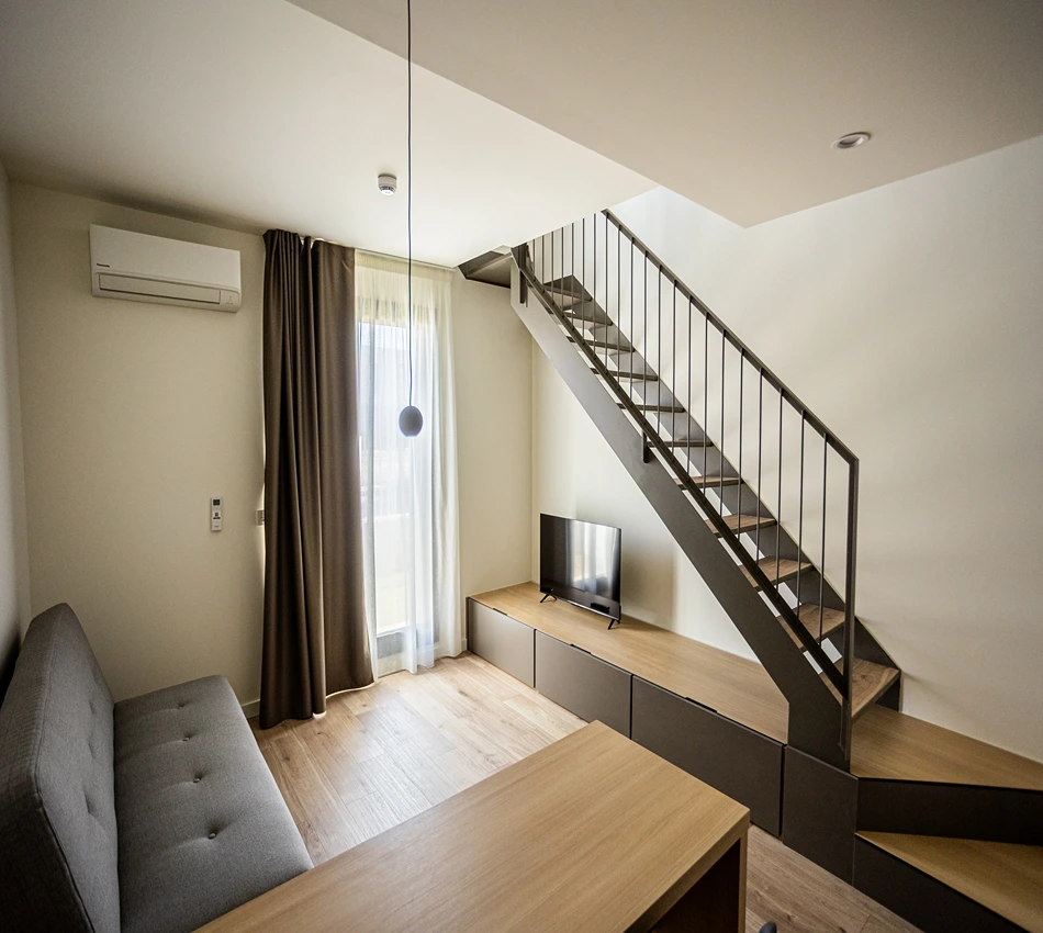 Sala moderna con escalera, sof&aacute;, televisor y ventana grande.