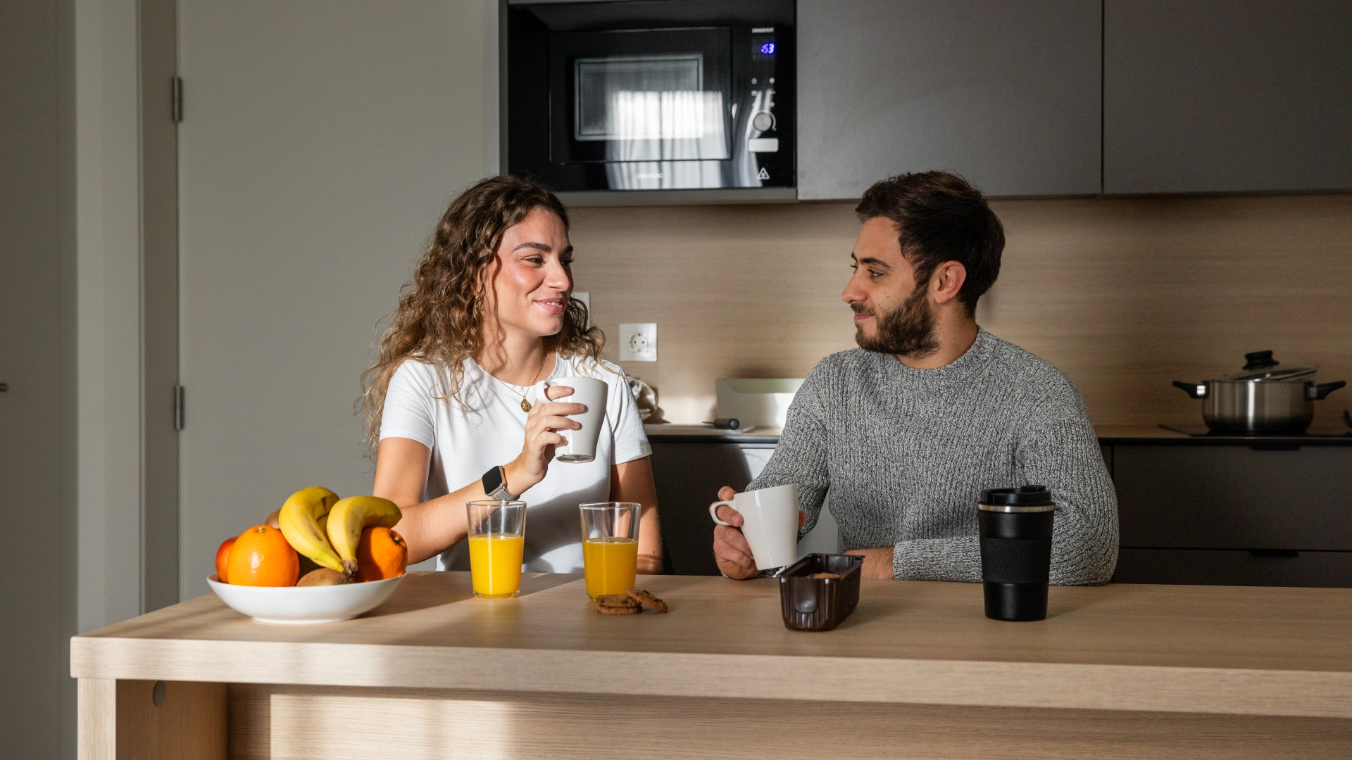 Dos personas conversan tomando caf&eacute; en una cocina con frutas y jugo.