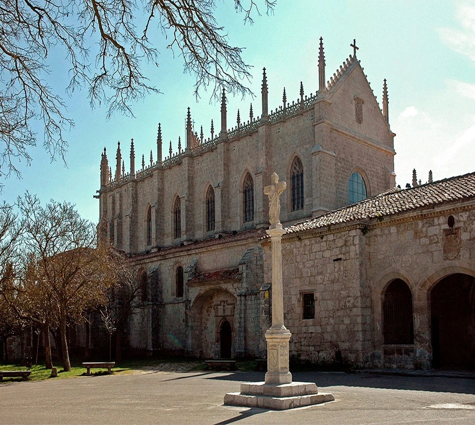 Edificio g&oacute;tico de piedra con cruz en primer plano y &aacute;rboles sin hojas.