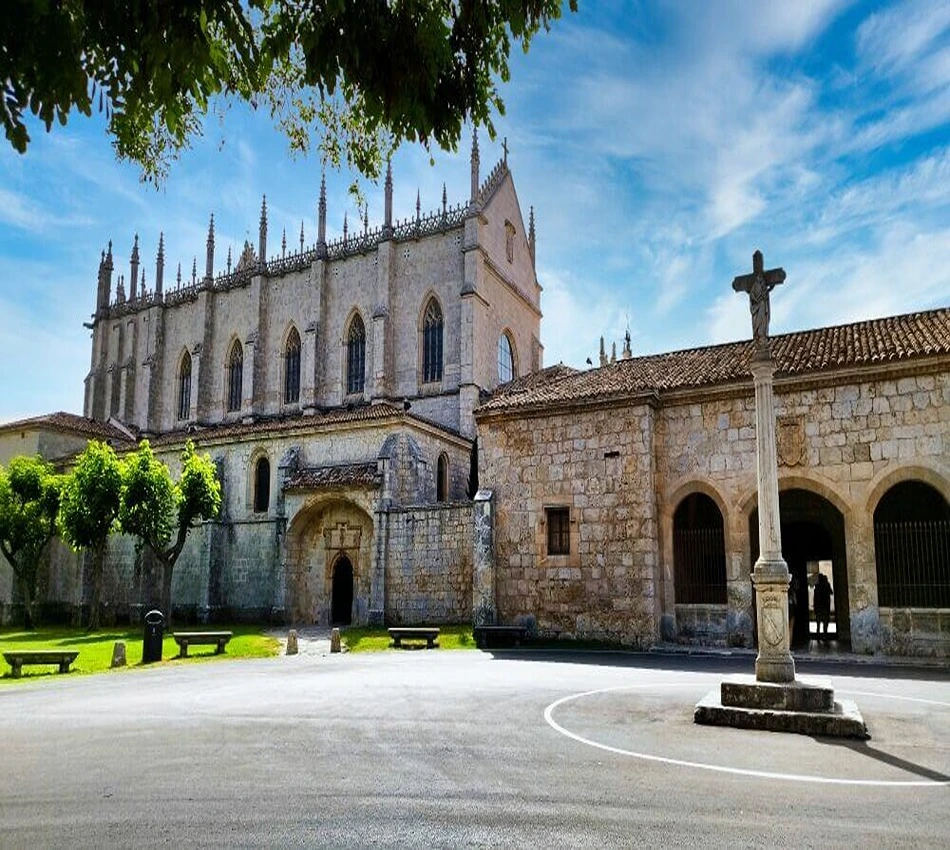 Edificio g&oacute;tico de piedra con cruz y &aacute;rboles en primer plano. Cielo despejado.