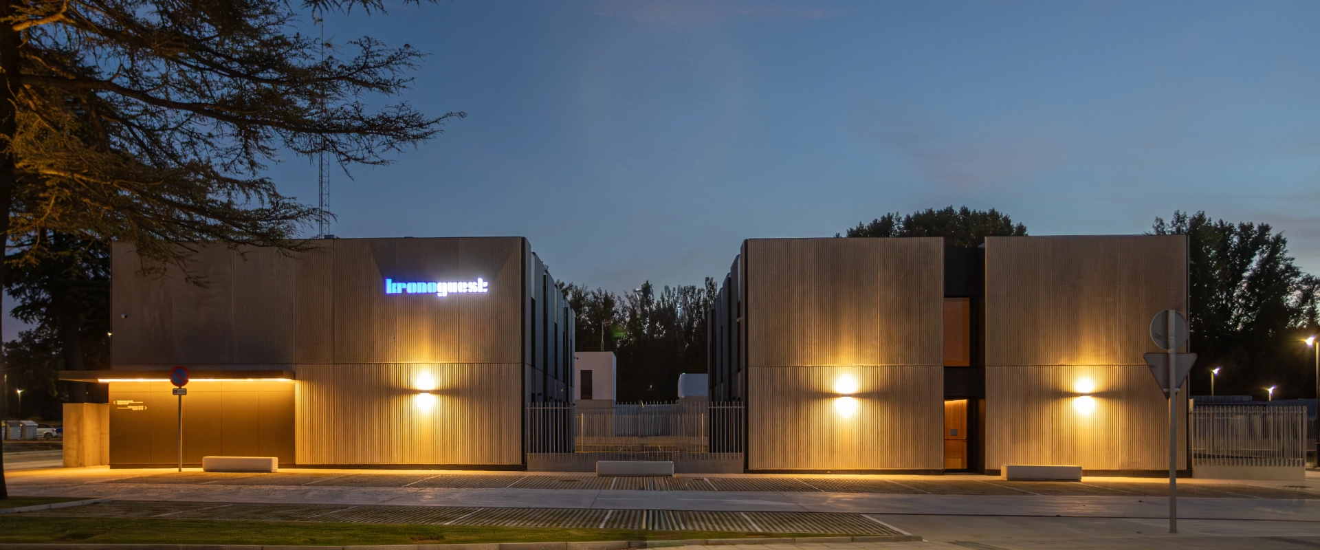 Edificio moderno iluminado al anochecer con un cielo azul y &aacute;rboles al fondo.