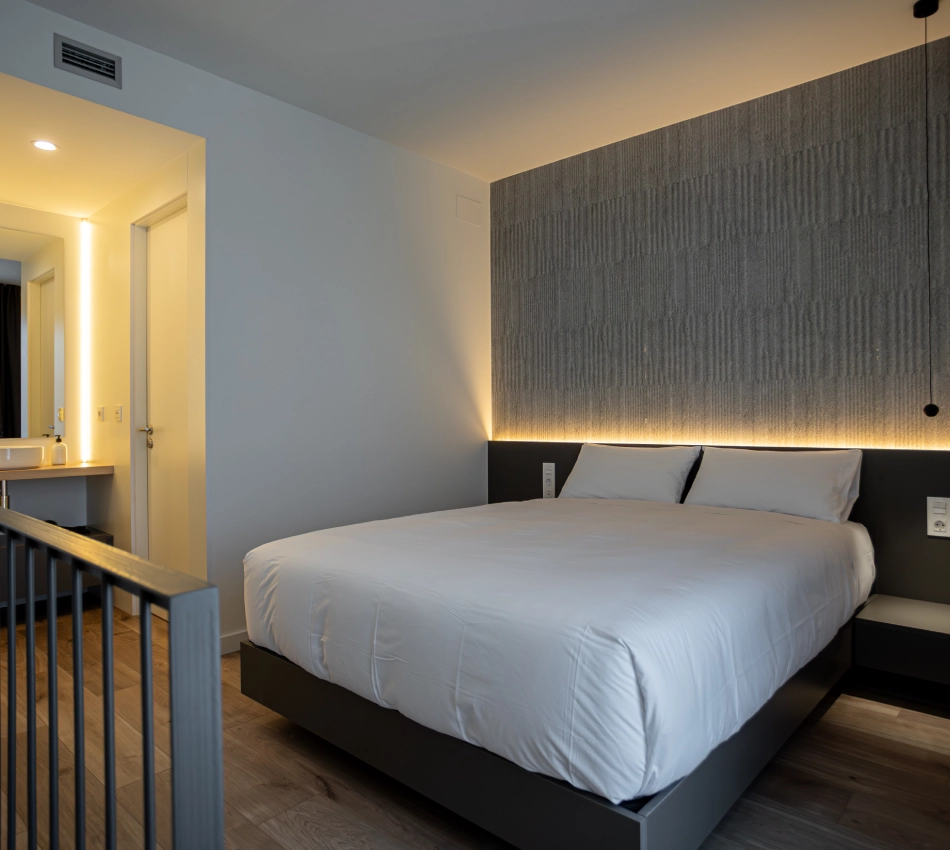 Dormitorio moderno con cama doble, luces c&aacute;lidas y ba&ntilde;o al fondo.