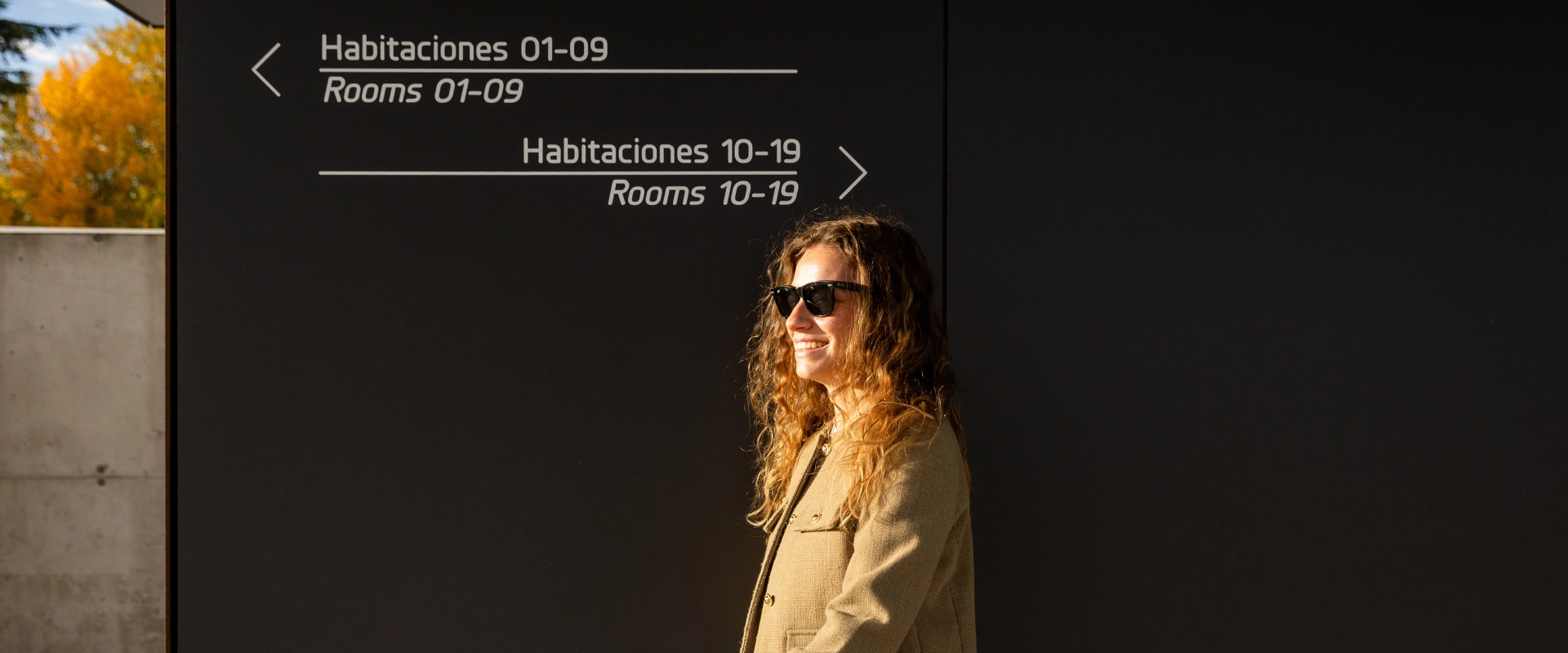 Persona sonriendo frente a un letrero de direcciones de habitaciones.