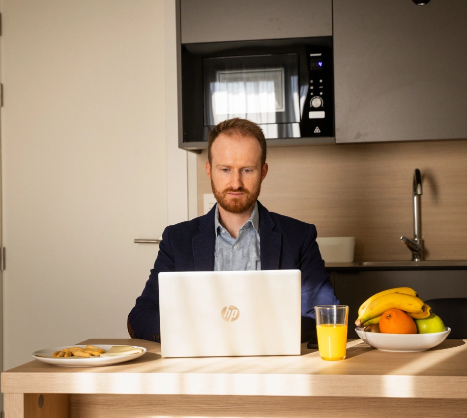 Hombre trabajando con laptop en una cocina, con frutas y jugo en la mesa.