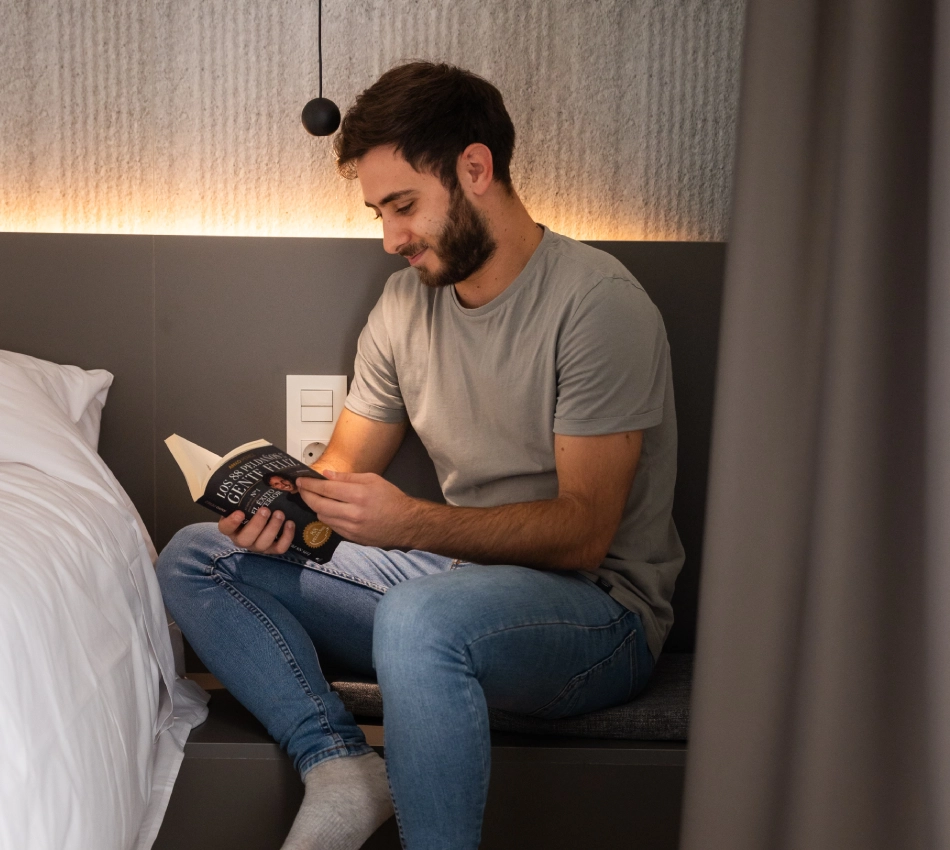 Hombre sentado en una cama leyendo un libro bajo una luz c&aacute;lida.